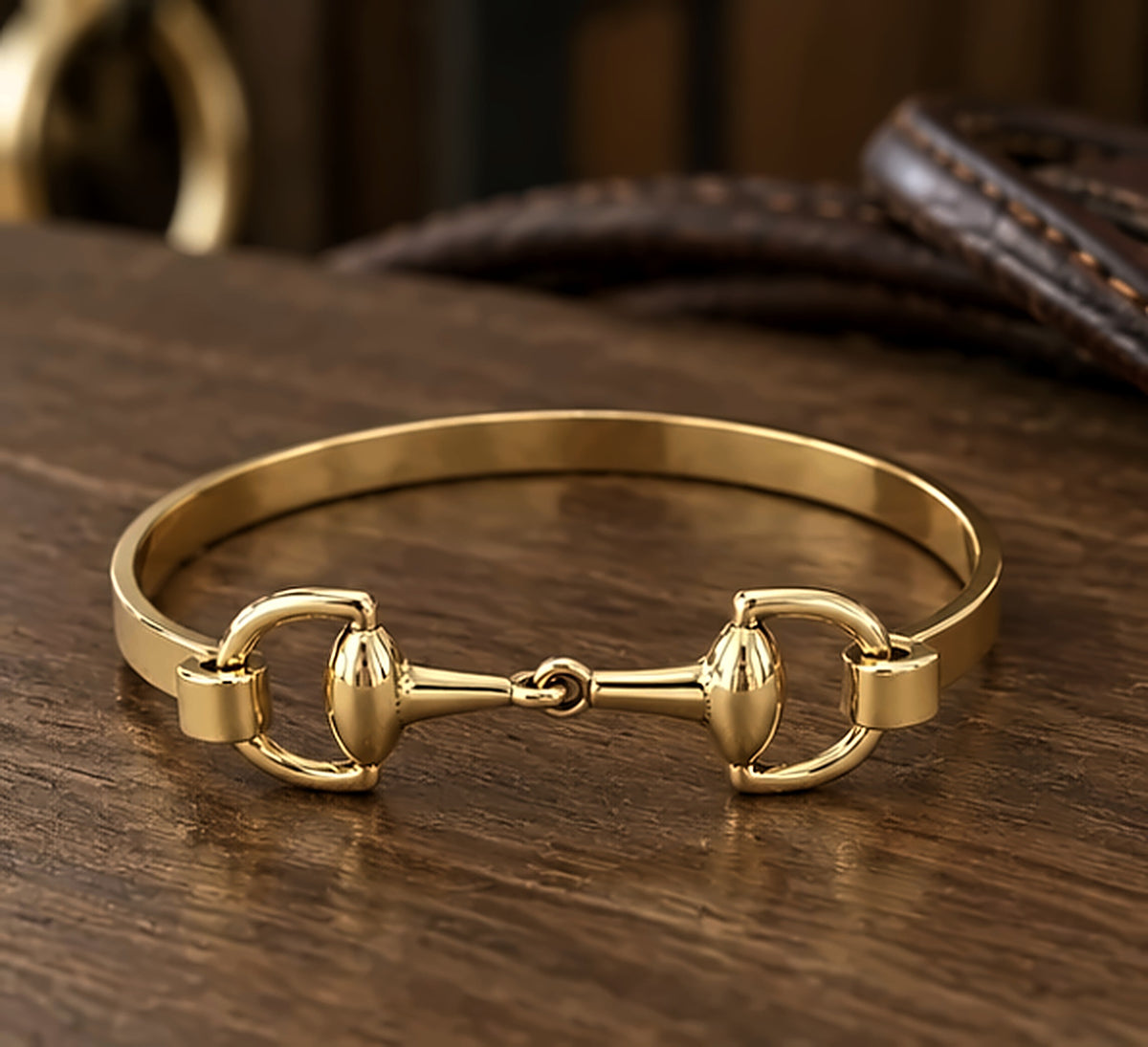 Gold snaffle bit bracelet on a wooden surface with leather reins behind it